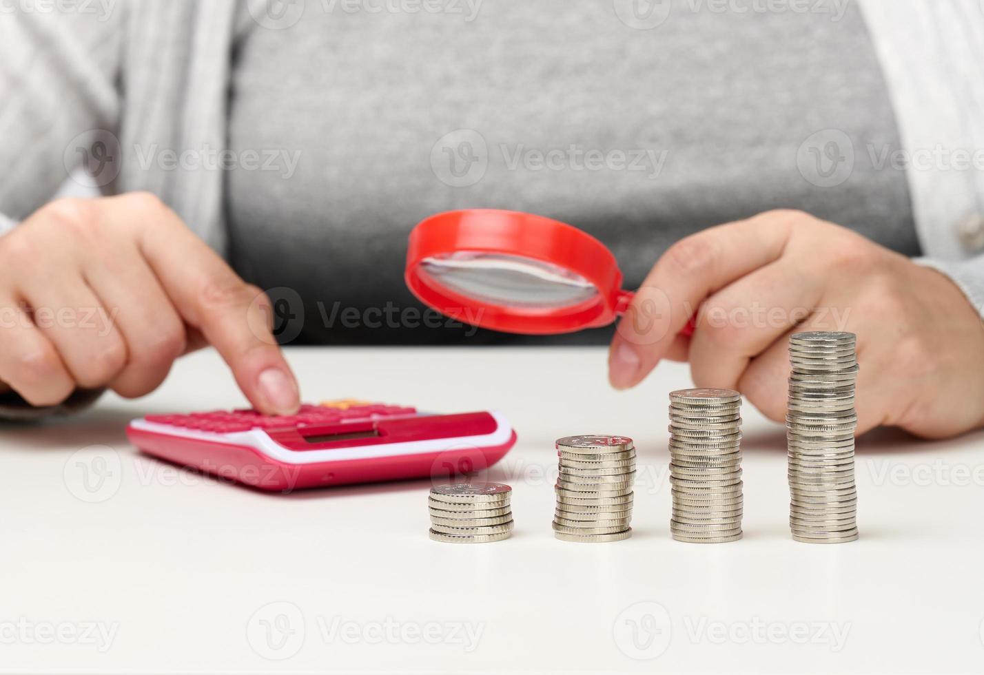 a woman sits at a table and counts on a calculator in the other hand a magnifying glass, a stack of coins on the table. Calculation of expenses and income photo