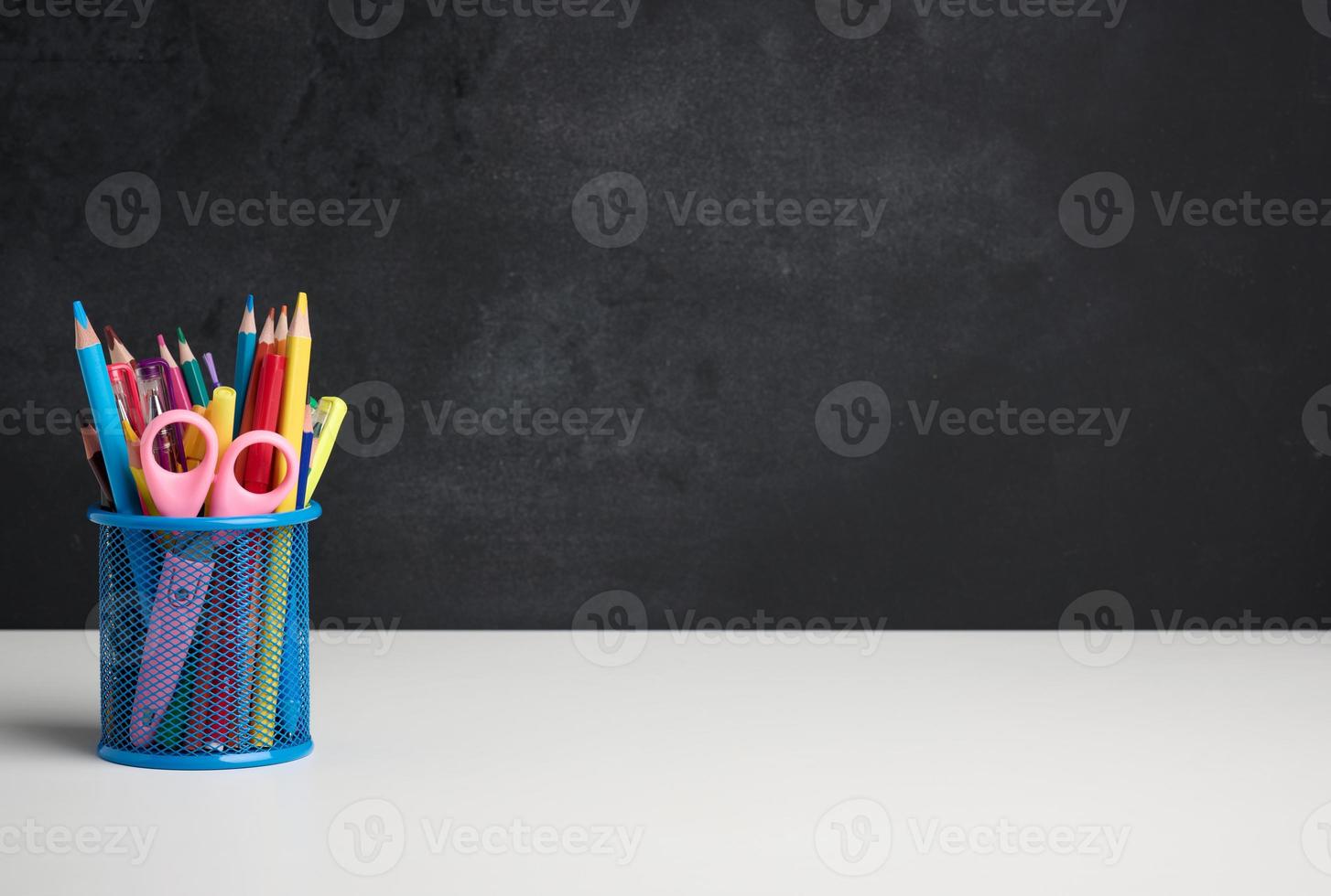 metal glass with pens, pencils and felt-tip pens and scissors on the background of an empty black chalk board, white table photo