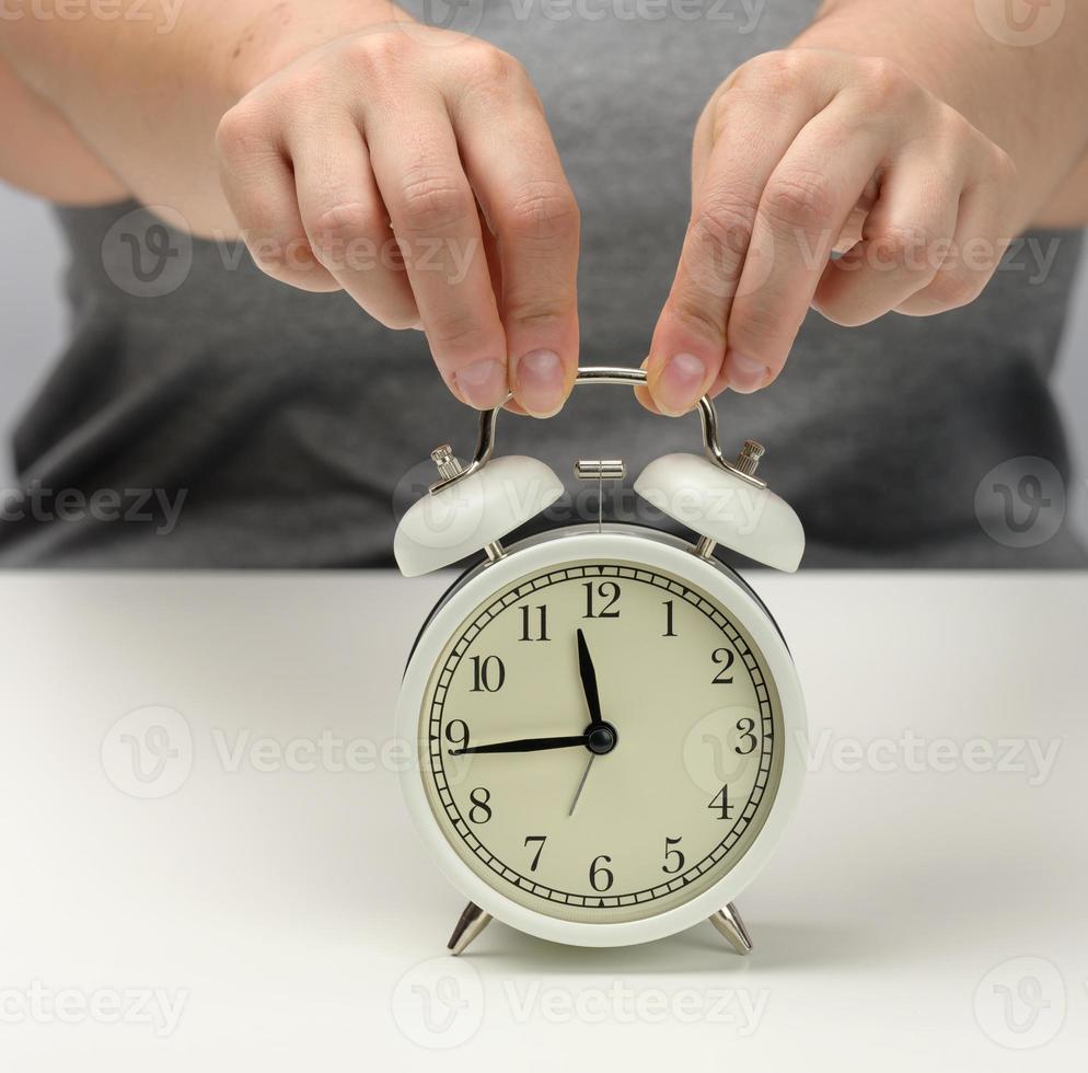 female hand holds a white metal alarm clock on a white table, the time is fifteen to twelve. Concept of the deadline for the assignment photo