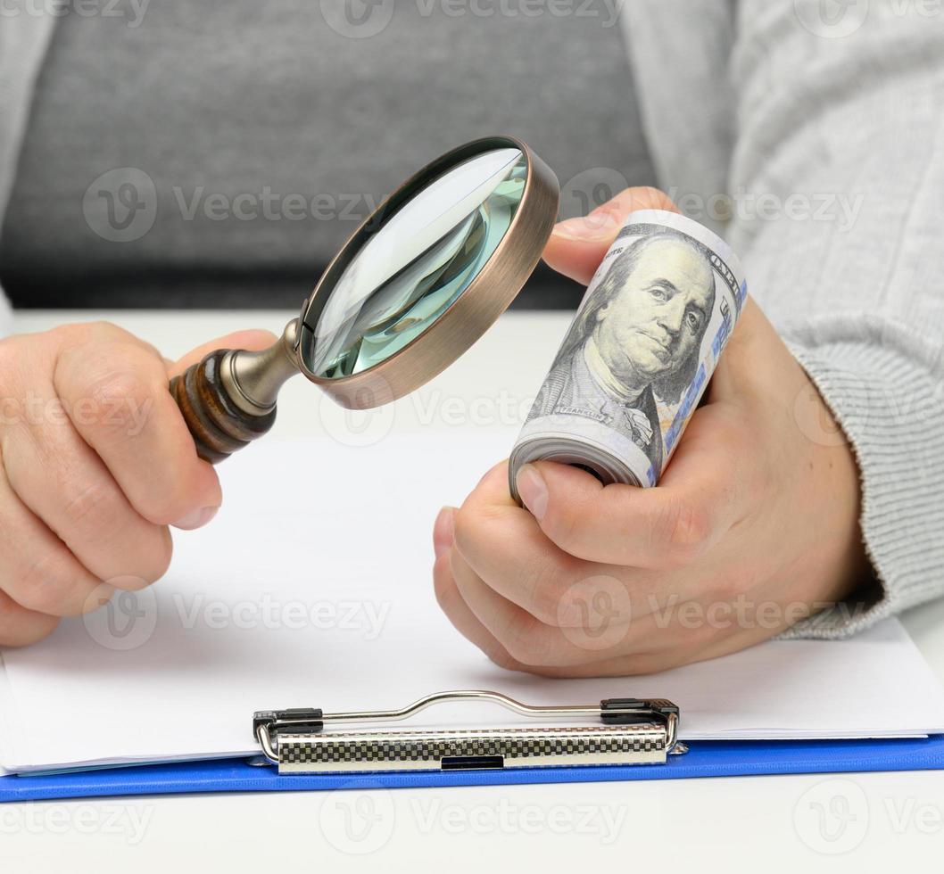 a stack of one hundred dollar bills in a woman's hand and a wooden magnifying glass on a white table. Budget analysis, income and expenses, profit calculation photo