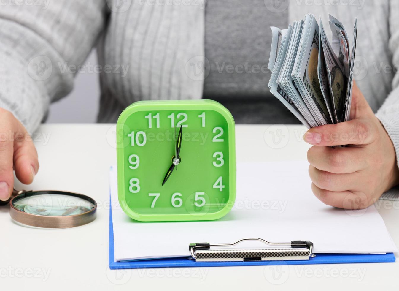 stack of one hundred dollar bills in a woman's hand and a wooden magnifying glass on a white table. Budget analysis, income and expenses photo