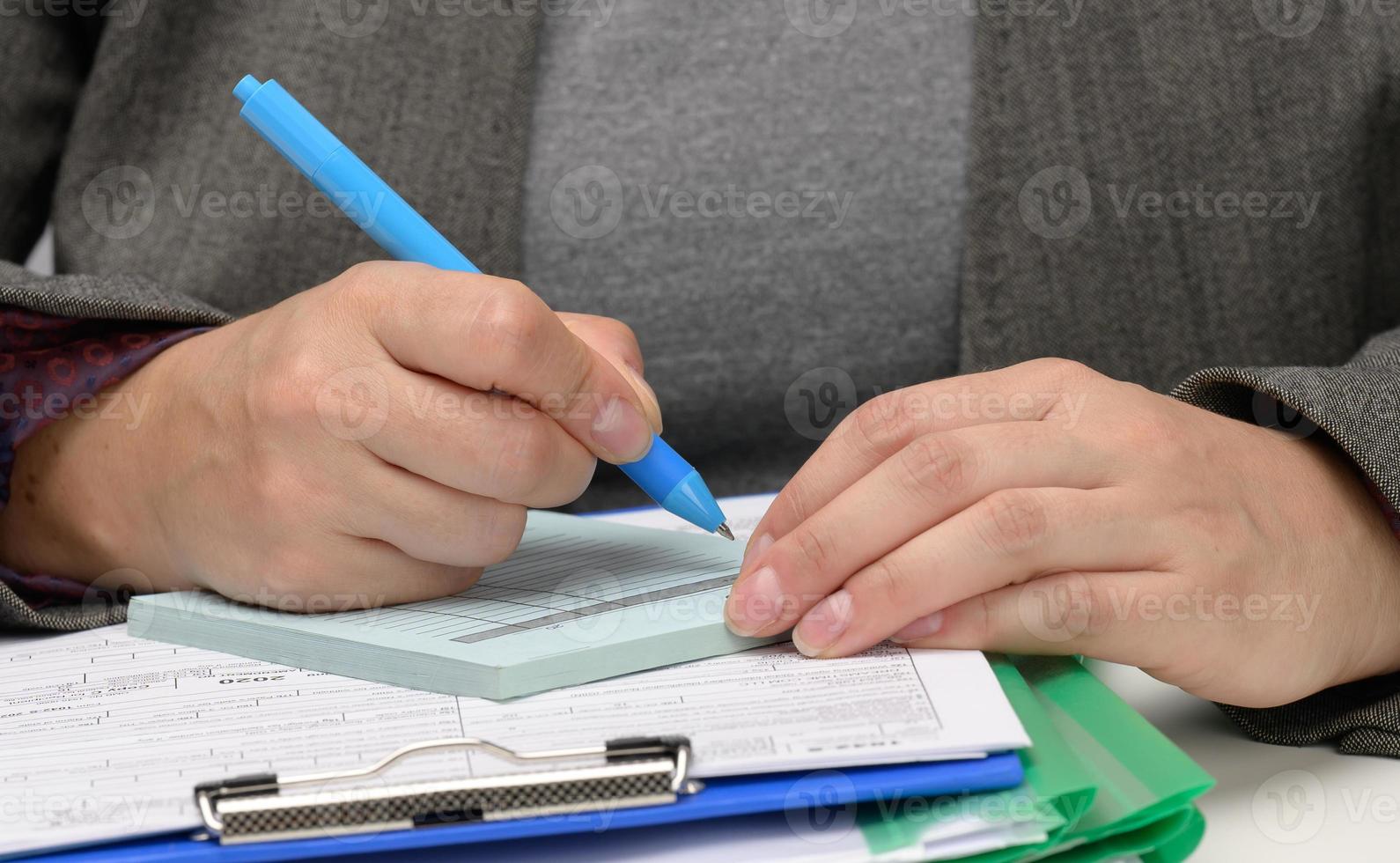 una mujer de apariencia caucásica se sienta con ropa gris en una mesa blanca y firma documentos con un bolígrafo de metal. empresario en el trabajo, firma en contrato y factura foto