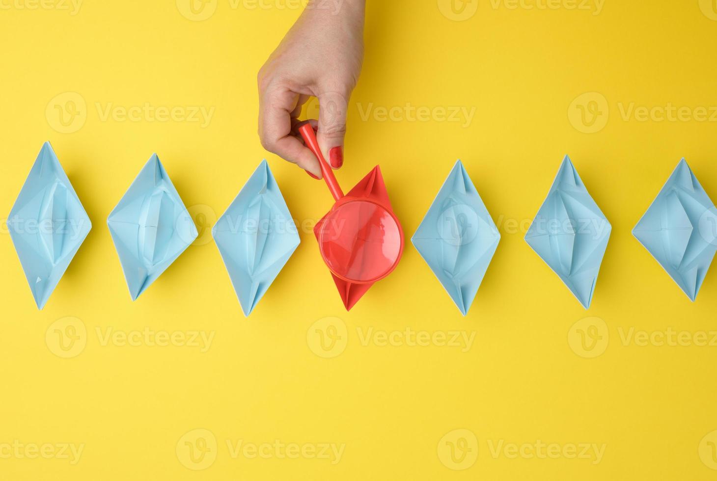 female hand holds a magnifying glass over a row of paper boats on a yellow background. Talent search concept photo