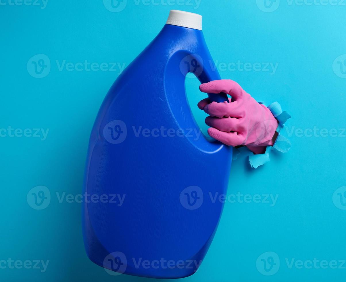 blue plastic bottle with liquid detergent in a female hand on a blue background. A part of the body sticks out of a torn hole in the background photo