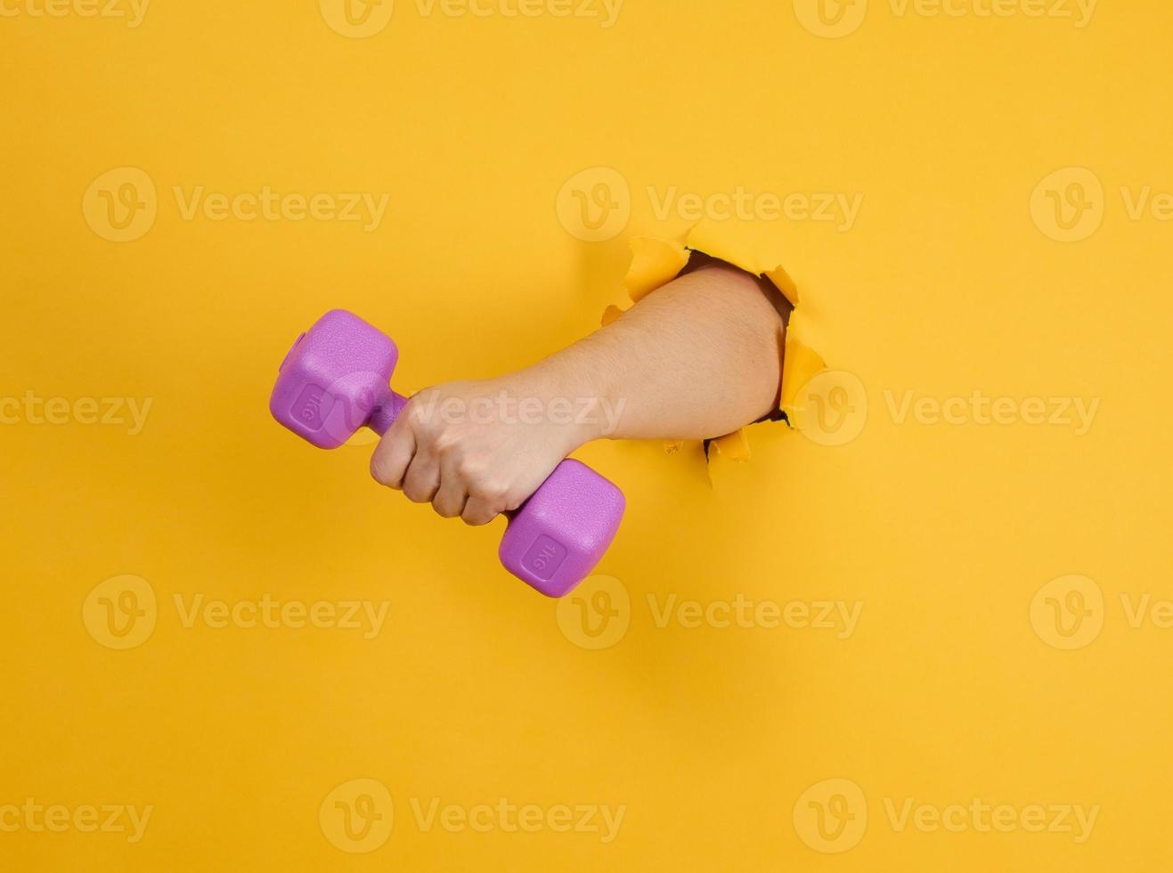 female hand holds a plastic kilogram dumbbell on a yellow background, a part of the body sticks out of a torn hole in a paper background. Healthy lifestyle, sports photo