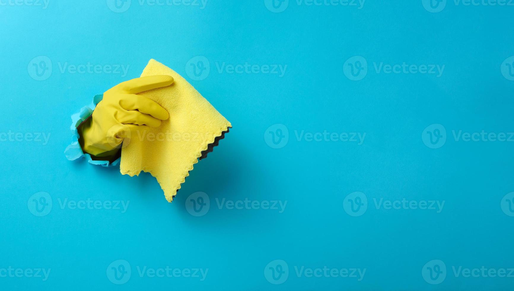 a hand in a yellow rubber cleaning glove holds a dry rag on a blue background. Part of the body stick out of the hole in the paper, banner photo