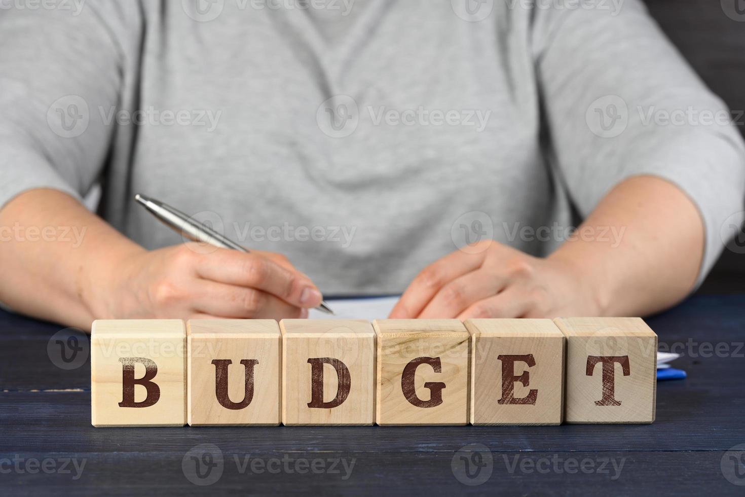 woman sits at the table and signs documents with a pen, in front of wooden cubes with the inscription budget photo