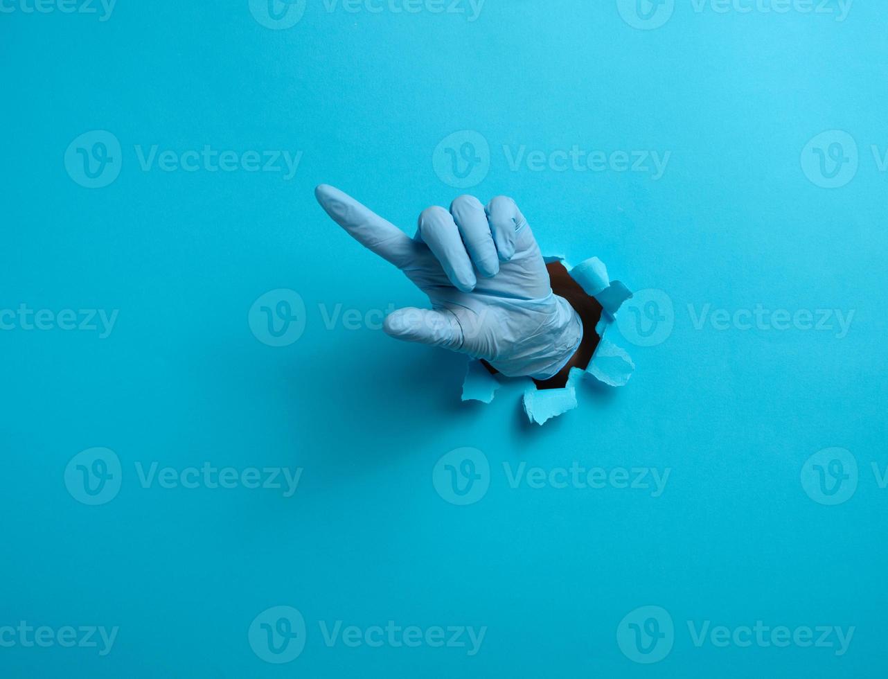 un mano en un azul médico guante es pega fuera de un Rasgado agujero en un azul papel fondo, el índice dedo es aumentó. parte de el cuerpo indica el dirección foto