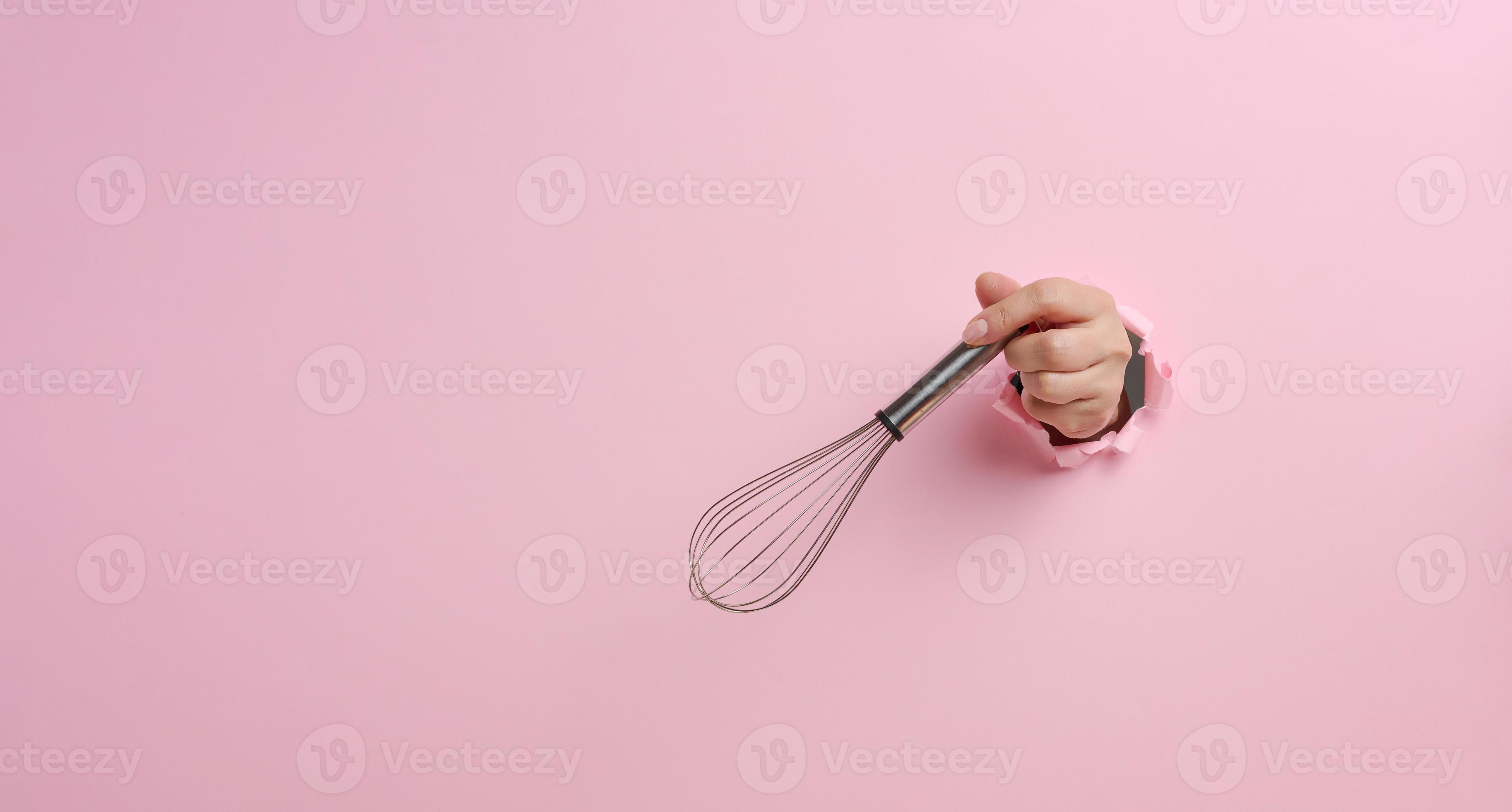 female hand holds a metal whisk for beating products on a pink