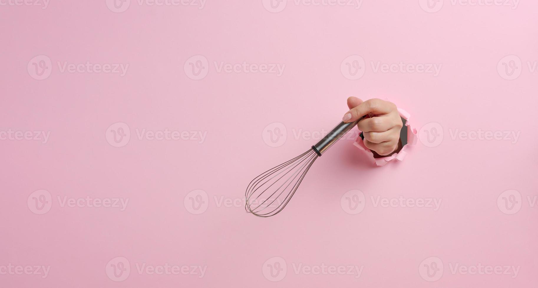 female hand holds a metal whisk for beating products on a pink background. A part of the body sticks out of a torn hole photo