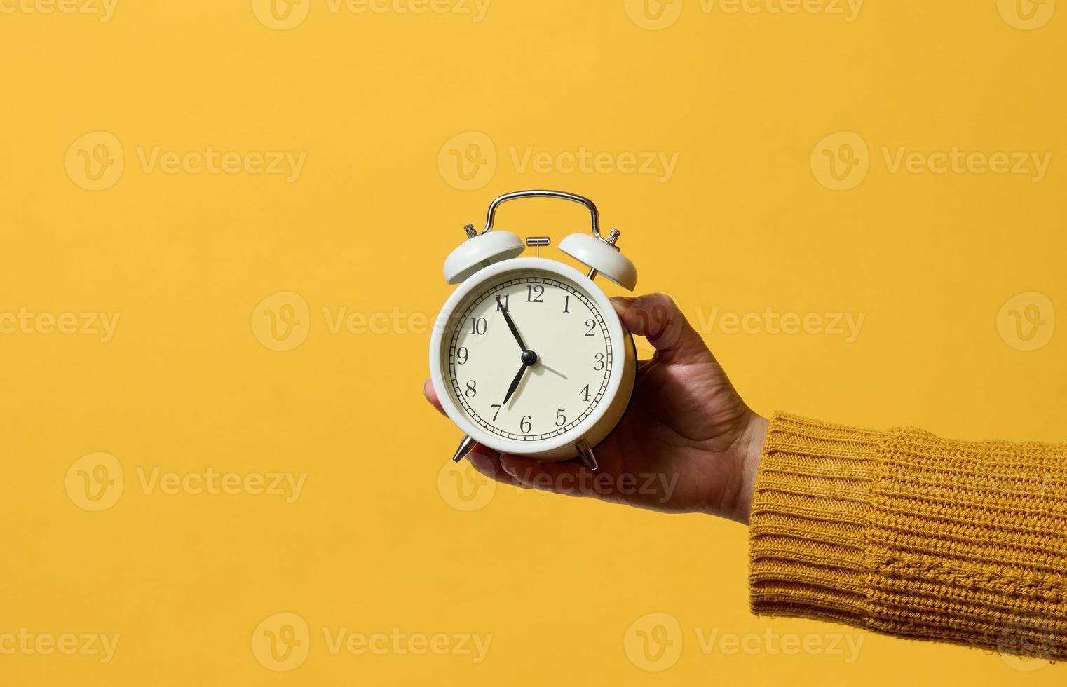 round alarm clock in a woman's hand, the time is five minutes to seven in the morning. Yellow background photo