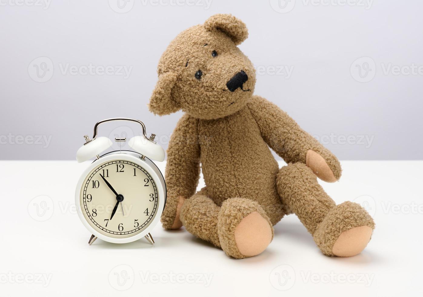 brown teddy bear sits on a white table and a round alarm clock, at five minutes to seven in the morning photo