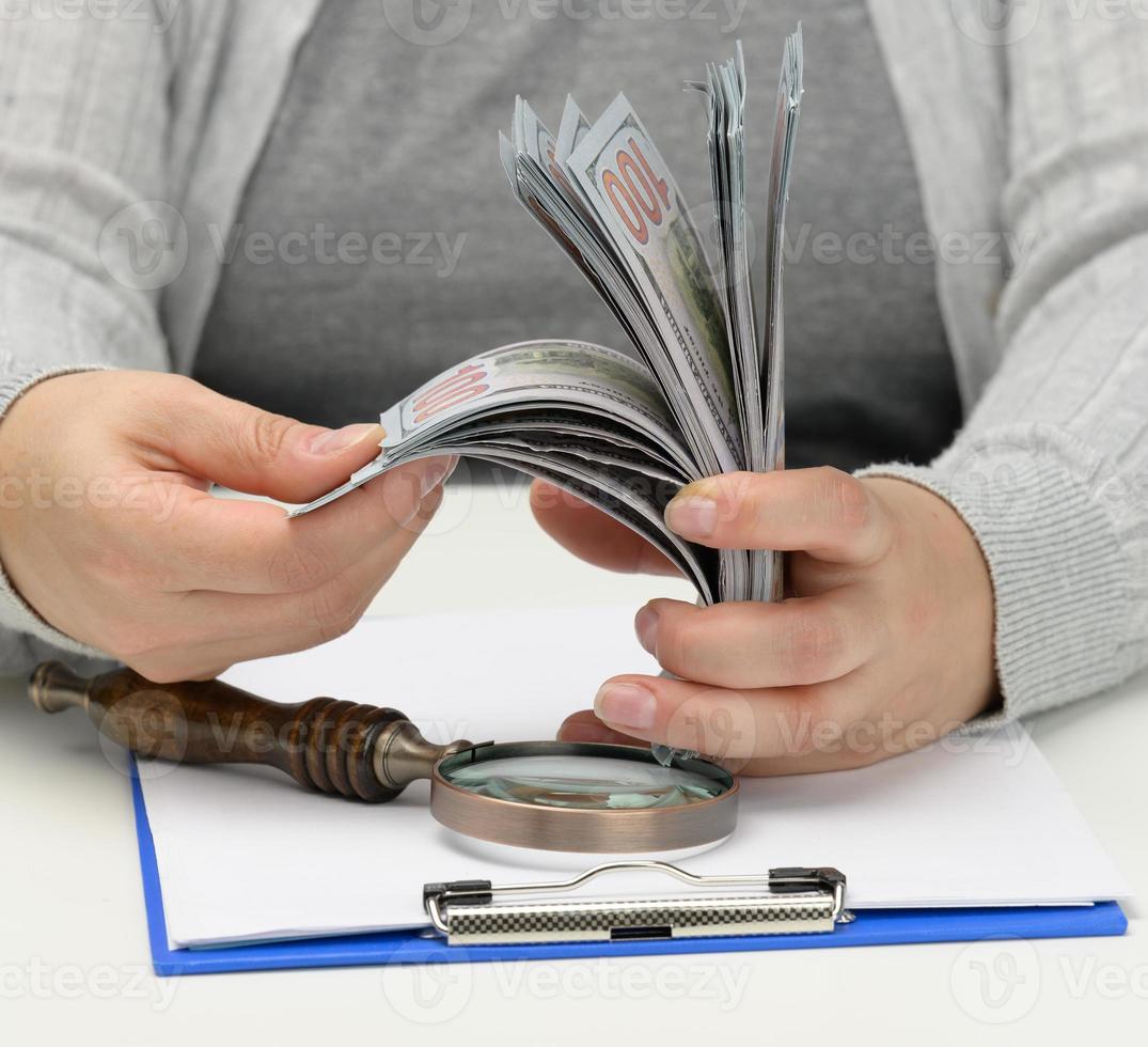 a stack of one hundred dollar bills in a woman's hand and a wooden magnifying glass on a white table. Budget analysis, income and expenses, problem finding photo