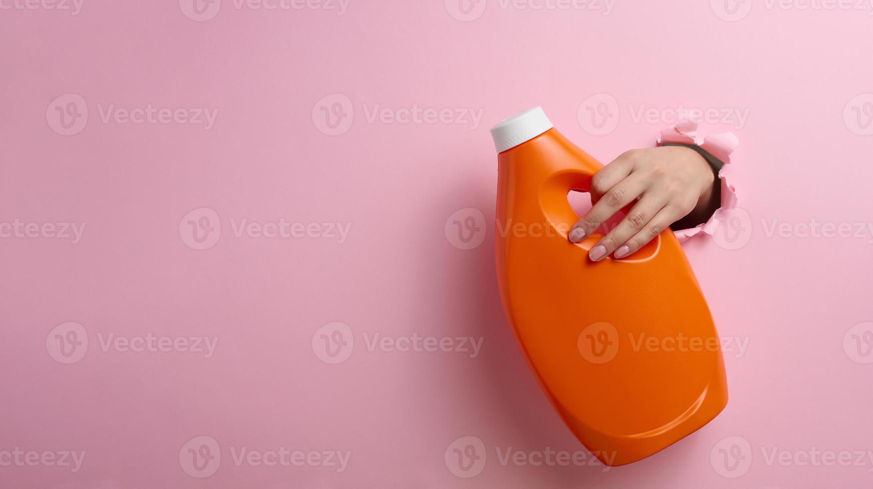 orange plastic bottle with liquid detergent in a female hand on a pink background. A part of the body sticks out of a torn hole in the background, a place for an inscription photo