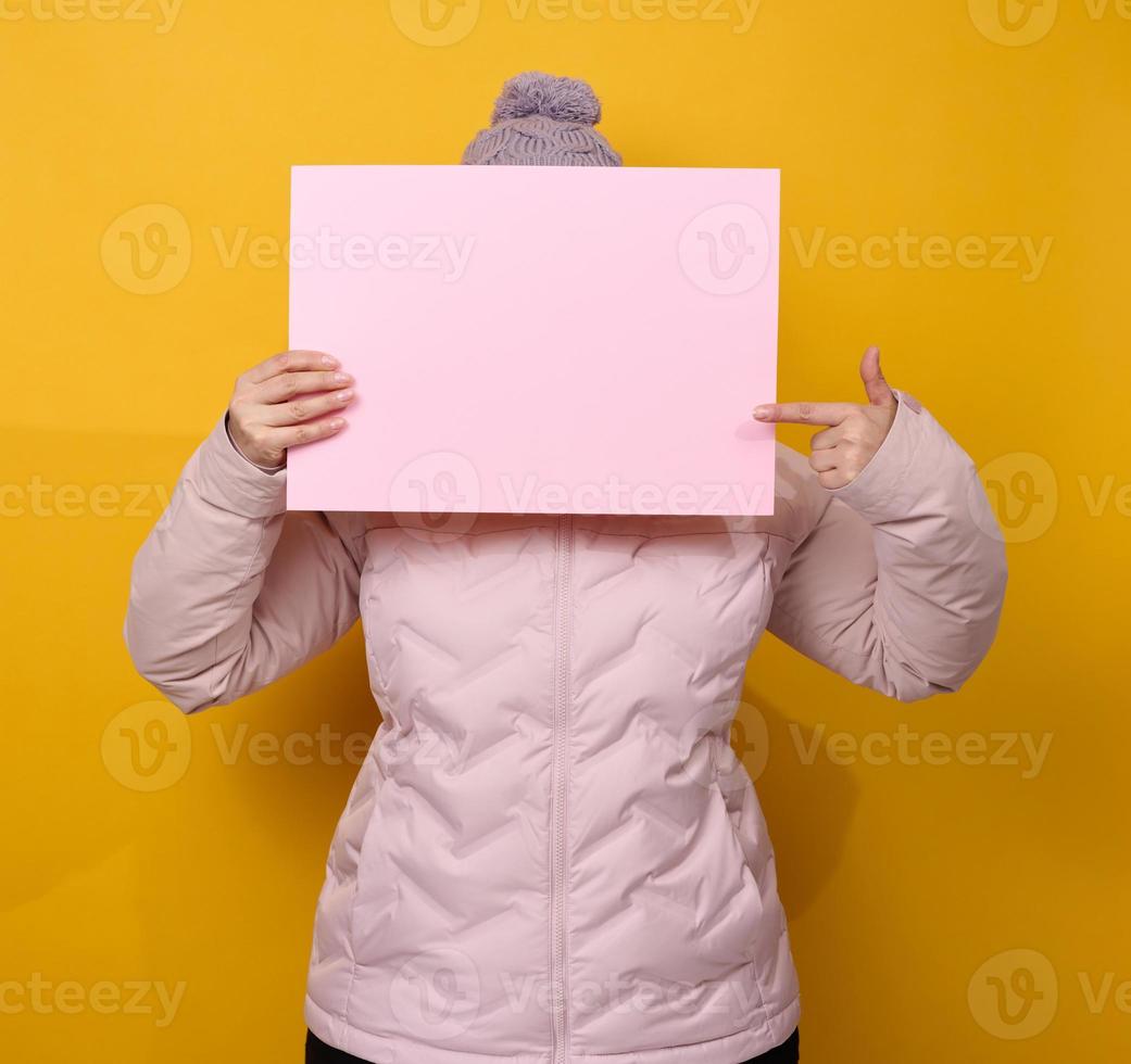 woman in pink winter jacket and hat holds blank pink sheet of paper on yellow background. Seasonal sale photo
