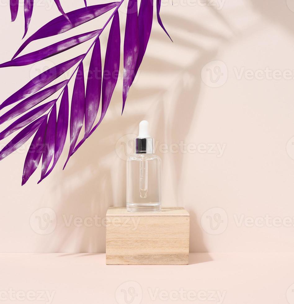 A white glass bottle with a pipette on a wooden podium and a purple palm leaf. photo