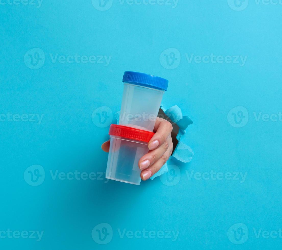 female hand holds an empty plastic jar for urine tests, the hand is sticking out of a torn hole in a blue background photo