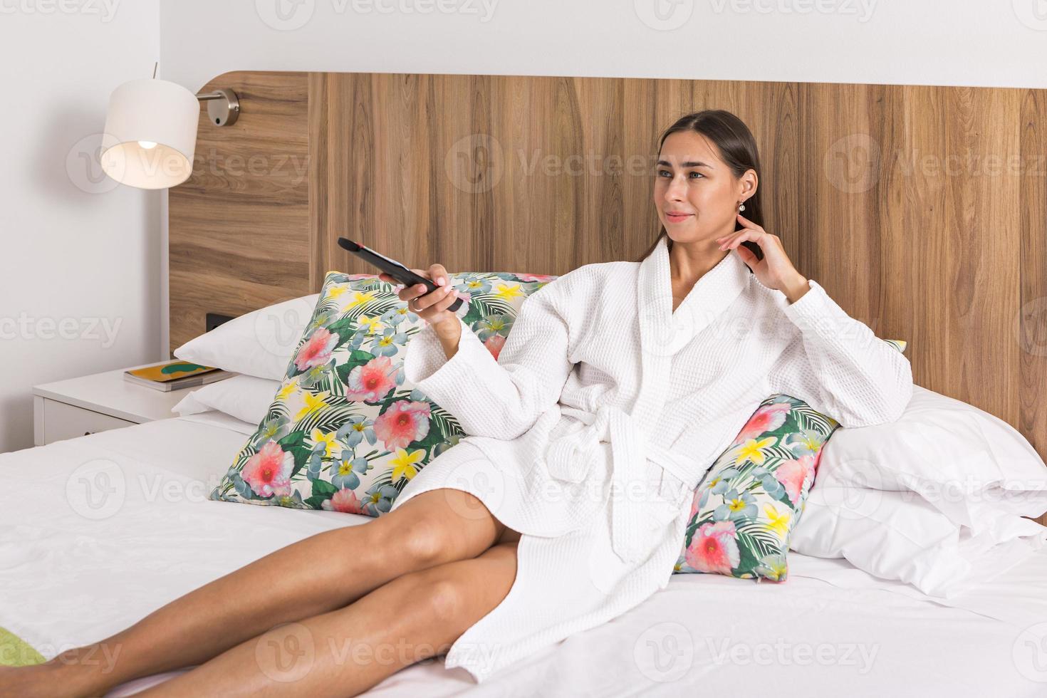mujer en bata de baño acostado en cama con remoto controlar en mano foto
