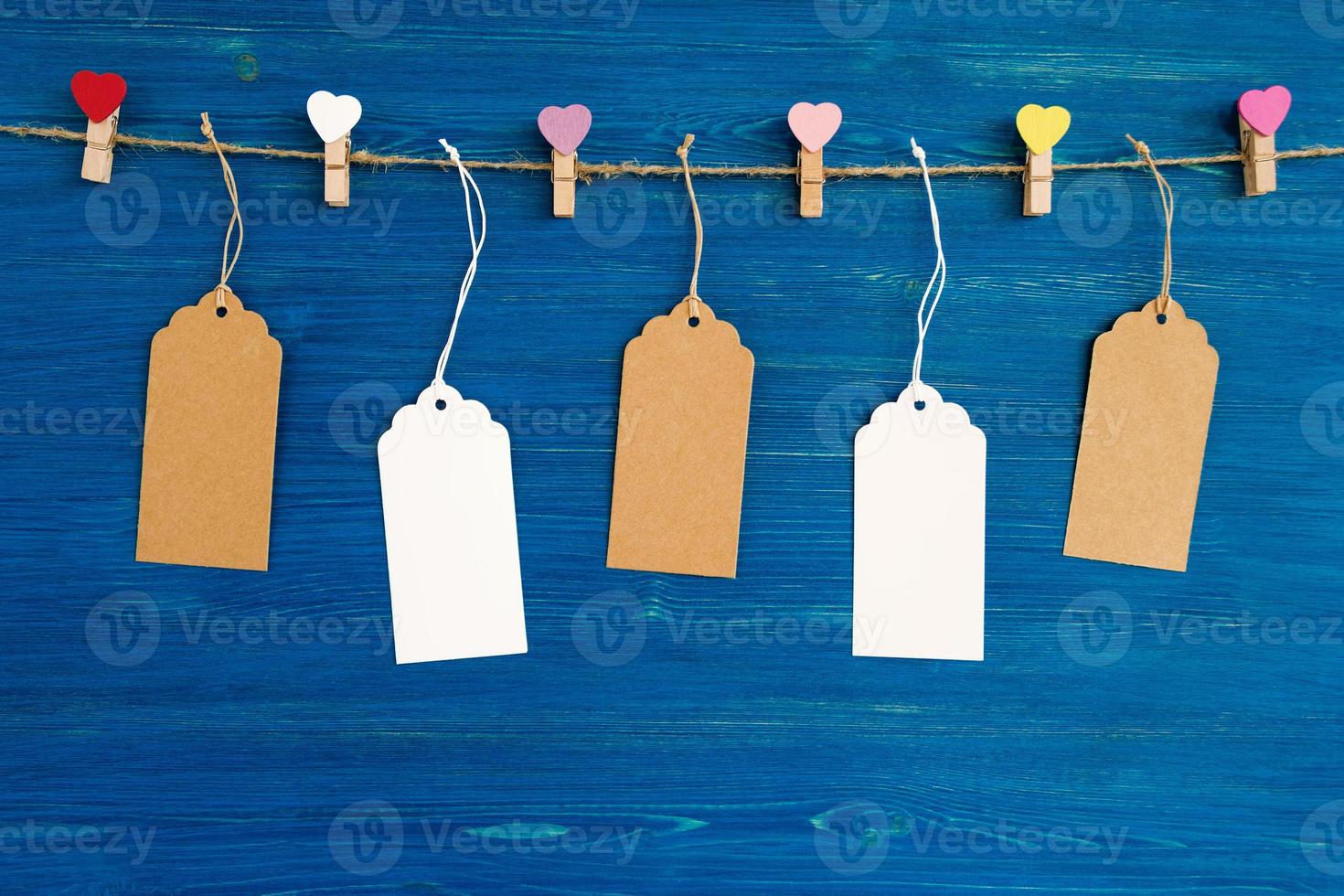 Brown and white blank paper price tags or labels set and wooden pins decorated on colored hearts hanging on a rope on the blue wooden background. photo