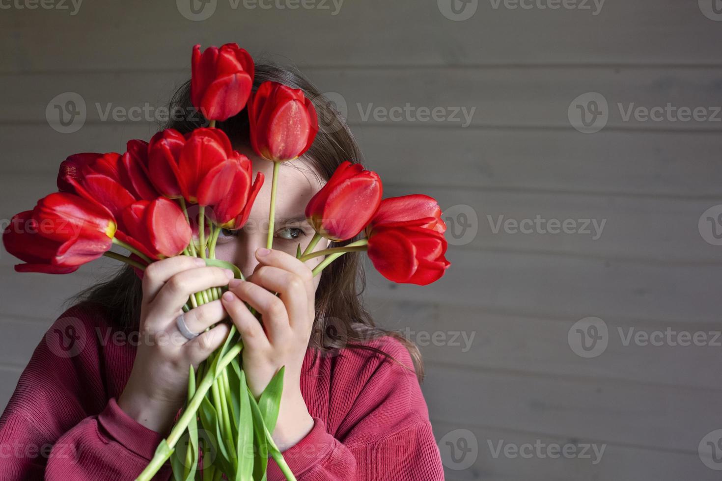 portrait of a brown-haired woman with a bouquet of red tulips on a gray wooden background. a girl in a red pullover with long hair holds a bouquet of tulips in her hands and fools around. Copy space. photo