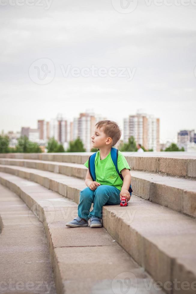 A boy is playing with a toy, sitting on the steps in the open air against the backdrop of skyscrapers and high-rise buildings. Journey. Lifestyle in the city. photo