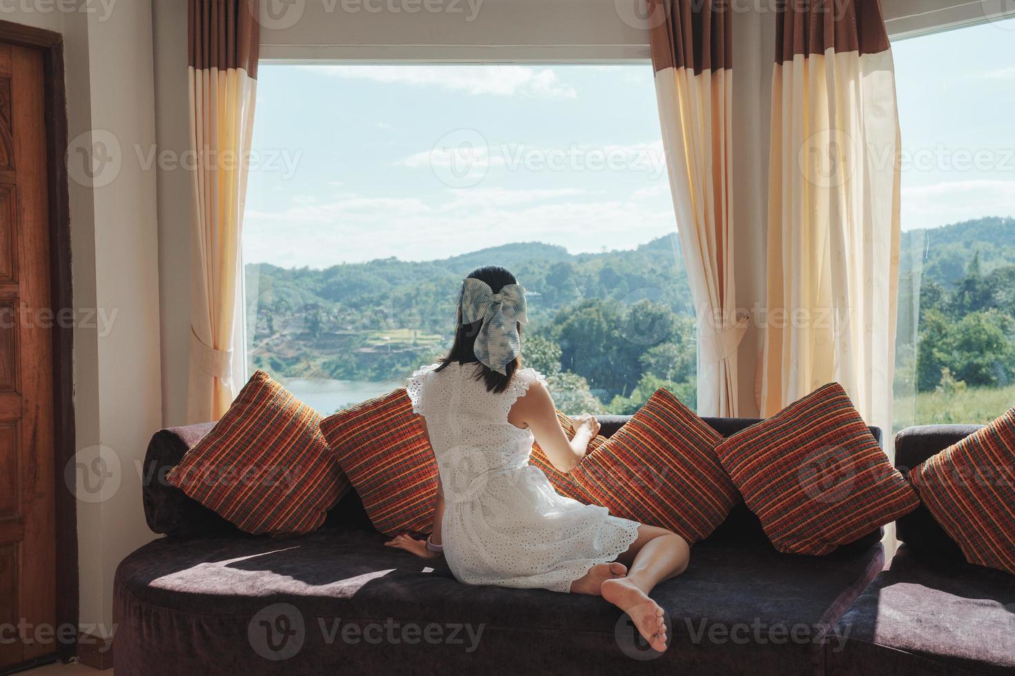 mujer asiática relajándose y disfrutando de la vista del bosque tropical en el sofá en la sala panorámica en un día brillante foto