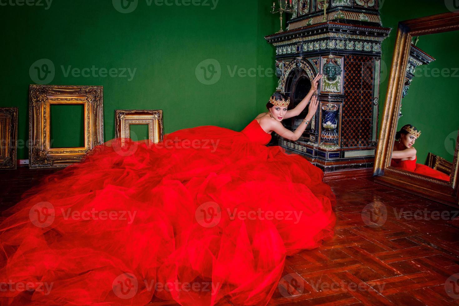 Beautiful woman in long red dress and in royal crown near fireplace in luxury interior in photo studio