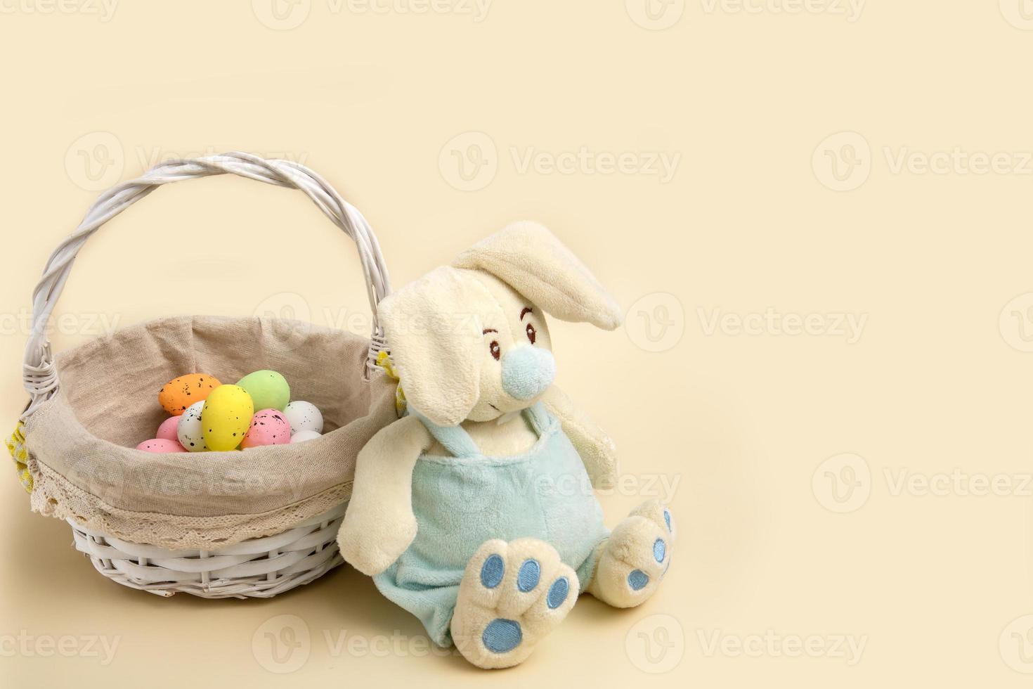 easter bunny sits by a basket with colorful eggs on a beige background with copy space photo