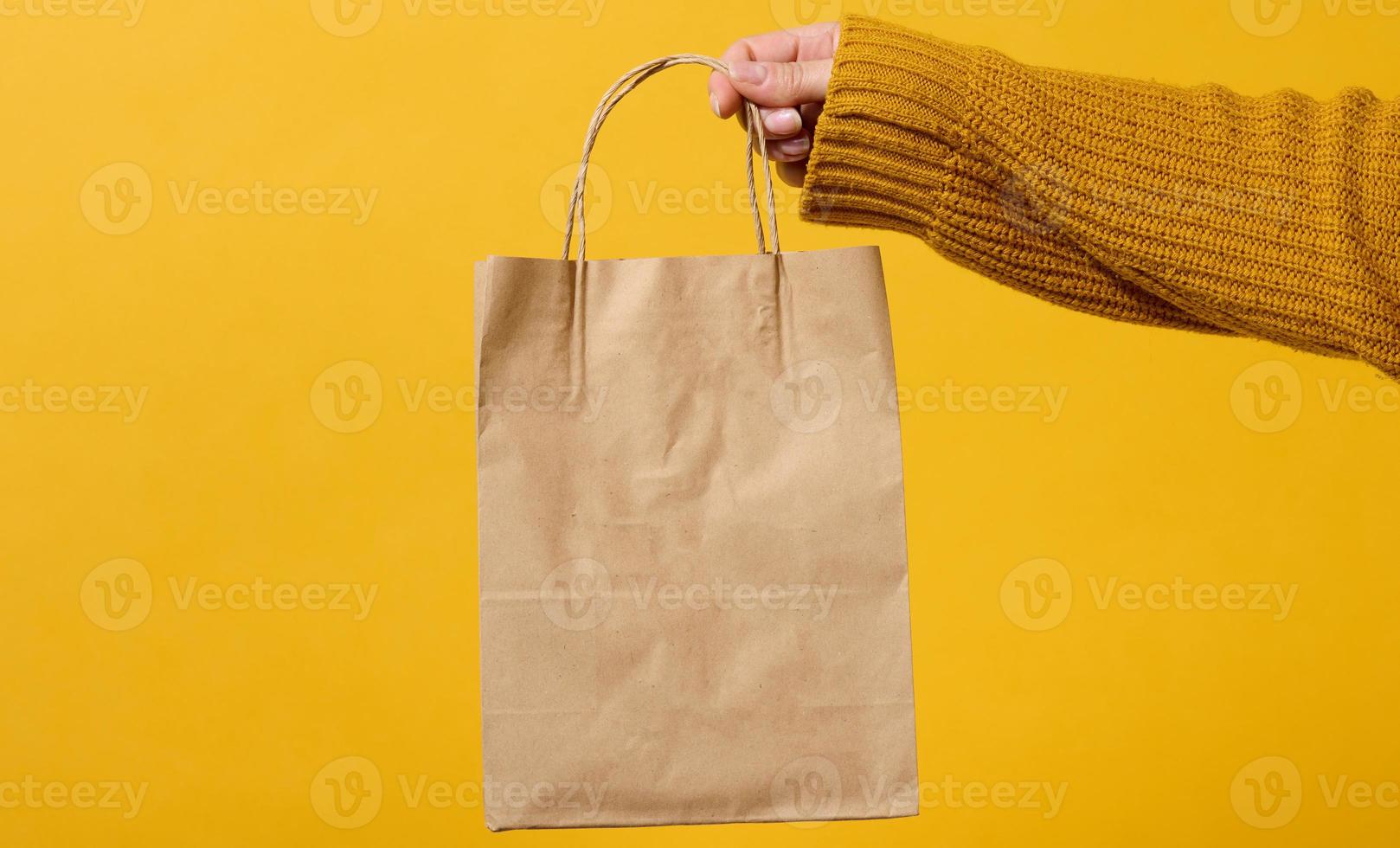 female hand in a sweater holds a brown craft paper bag with handles on a yellow background. Zero waste photo