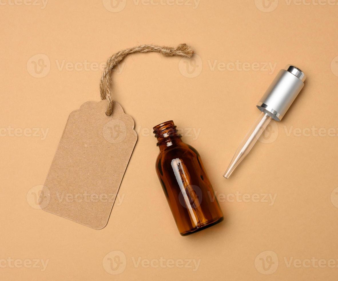 paper tag and glass cosmetic brown bottle with a pipette on a beige background. Cosmetics SPA branding mockup, top view photo