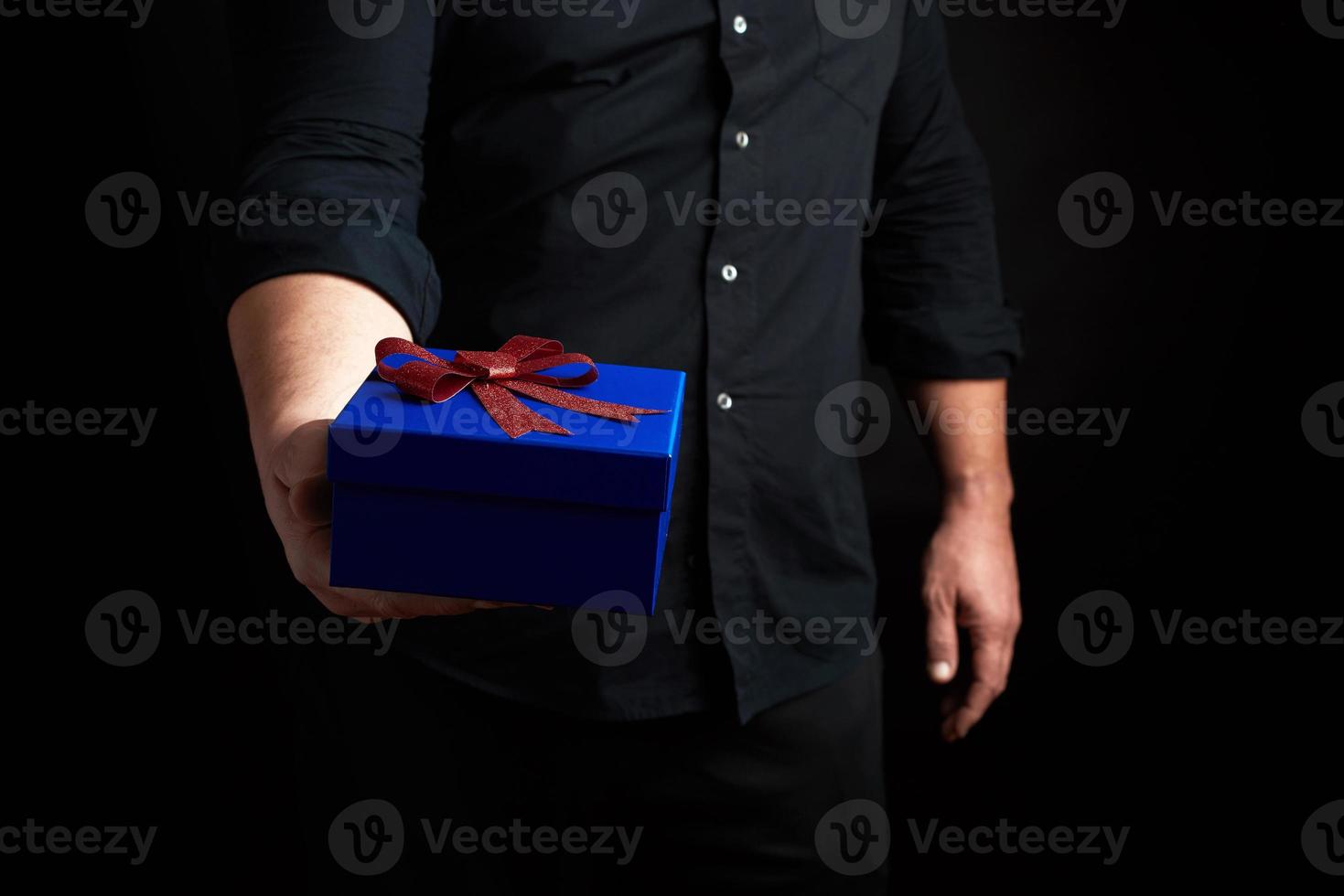 adulto hombre en un negro camisa sostiene un azul cuadrado caja con un rojo arco atado en un oscuro antecedentes foto