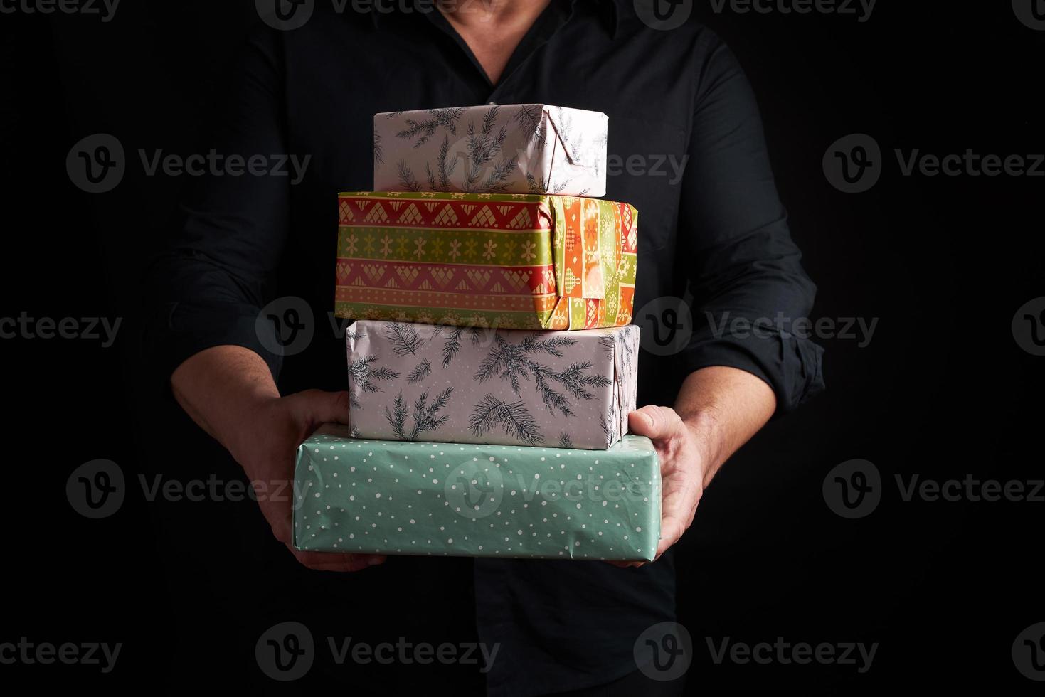 adult man in a black shirt holds in his hands a stack of paper-wrapped gifts on a black background photo