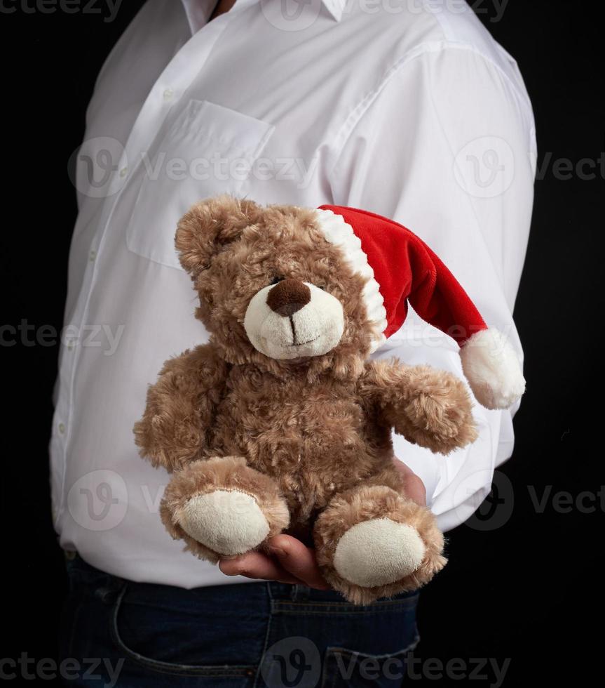 man in a white shirt holds a teddy brown bear in a red cap photo