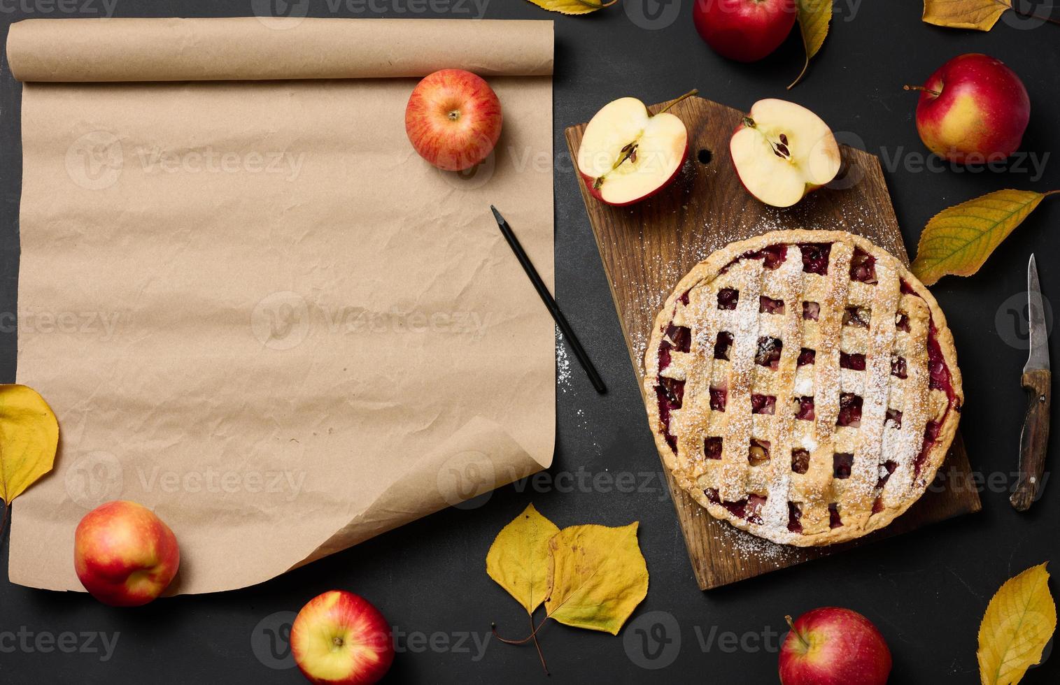 baked round traditional apple pie on a brown wooden board and fresh red apples. Nearby is a roll of brown paper for writing a recipe or menu photo