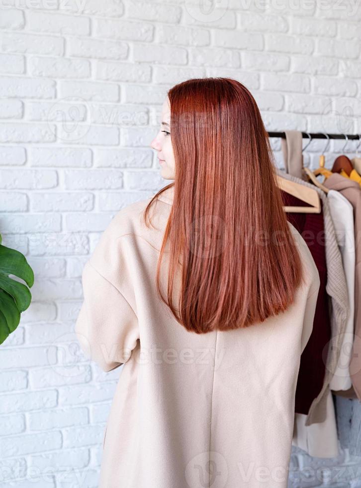 young beautiful woman wearing beige pastel coat, standing near clothes rack. Wardrobe change photo