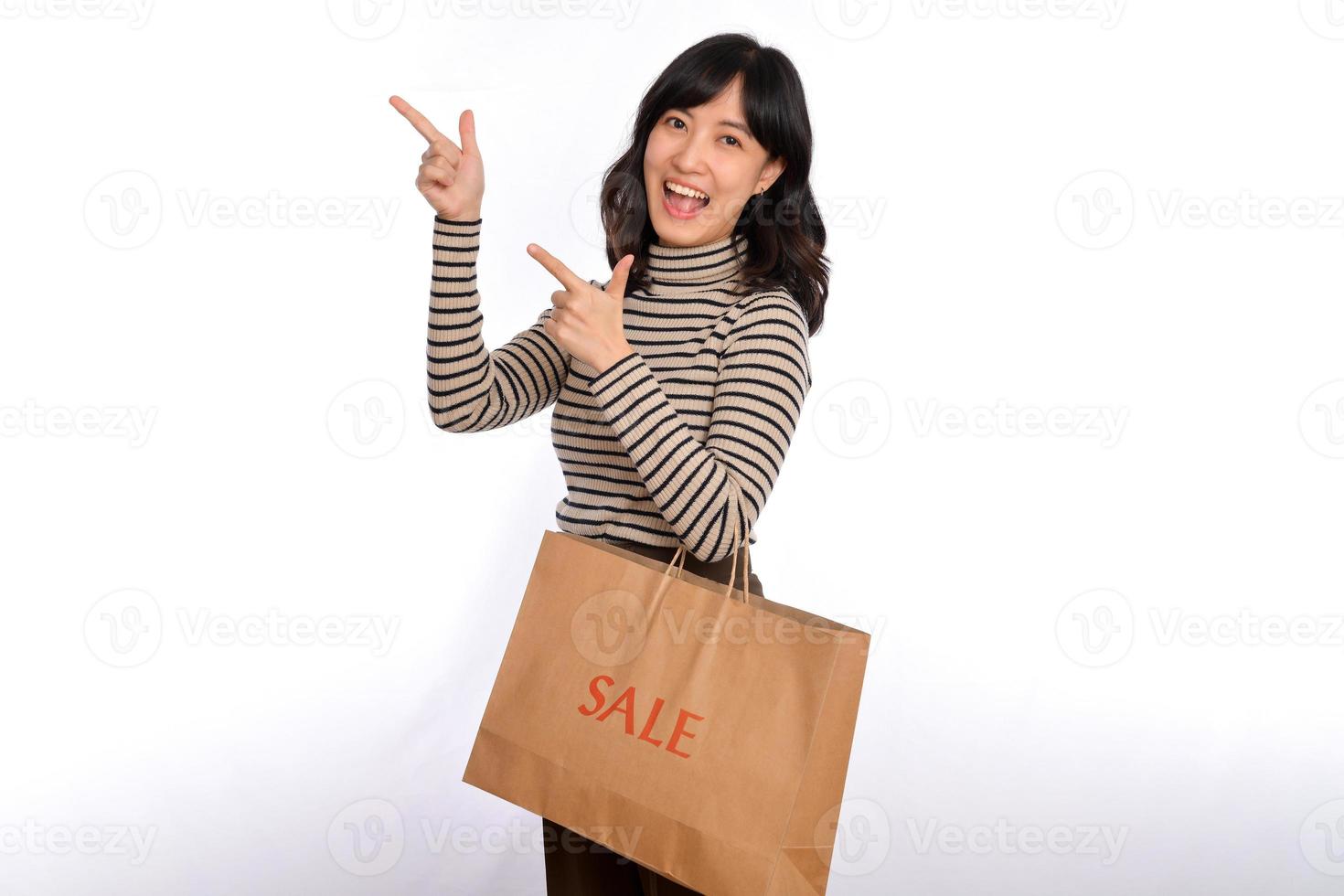 Beautiful young Asian woman on sweater cloth pointing up to copy space and looking at camera with smile face and sale shopping paper bag isolated on white background photo