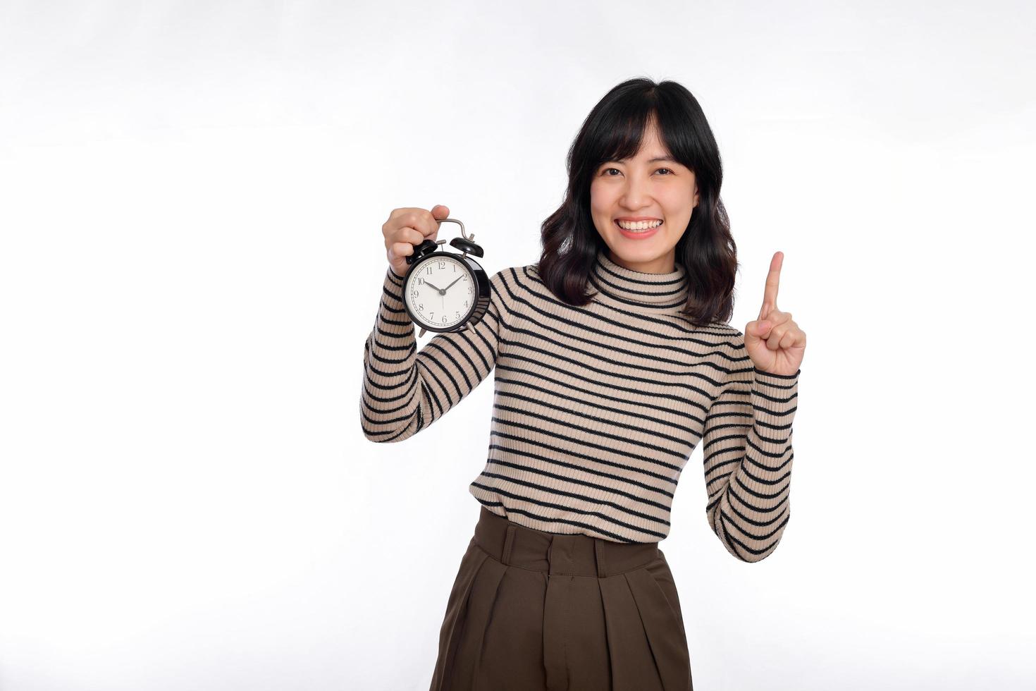 Smiling cheerful attractive young asian woman wearing sweater shirt holding alarm clock, point finger above and looking camera isolated on white background, studio portrait photo