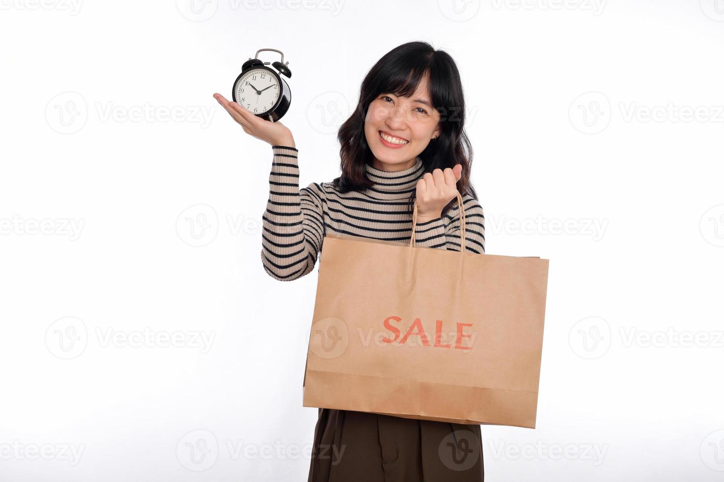 Portrait of happy young Asian woman with sweater shirt holding alarm clock and shopping paper back isolated on white background photo