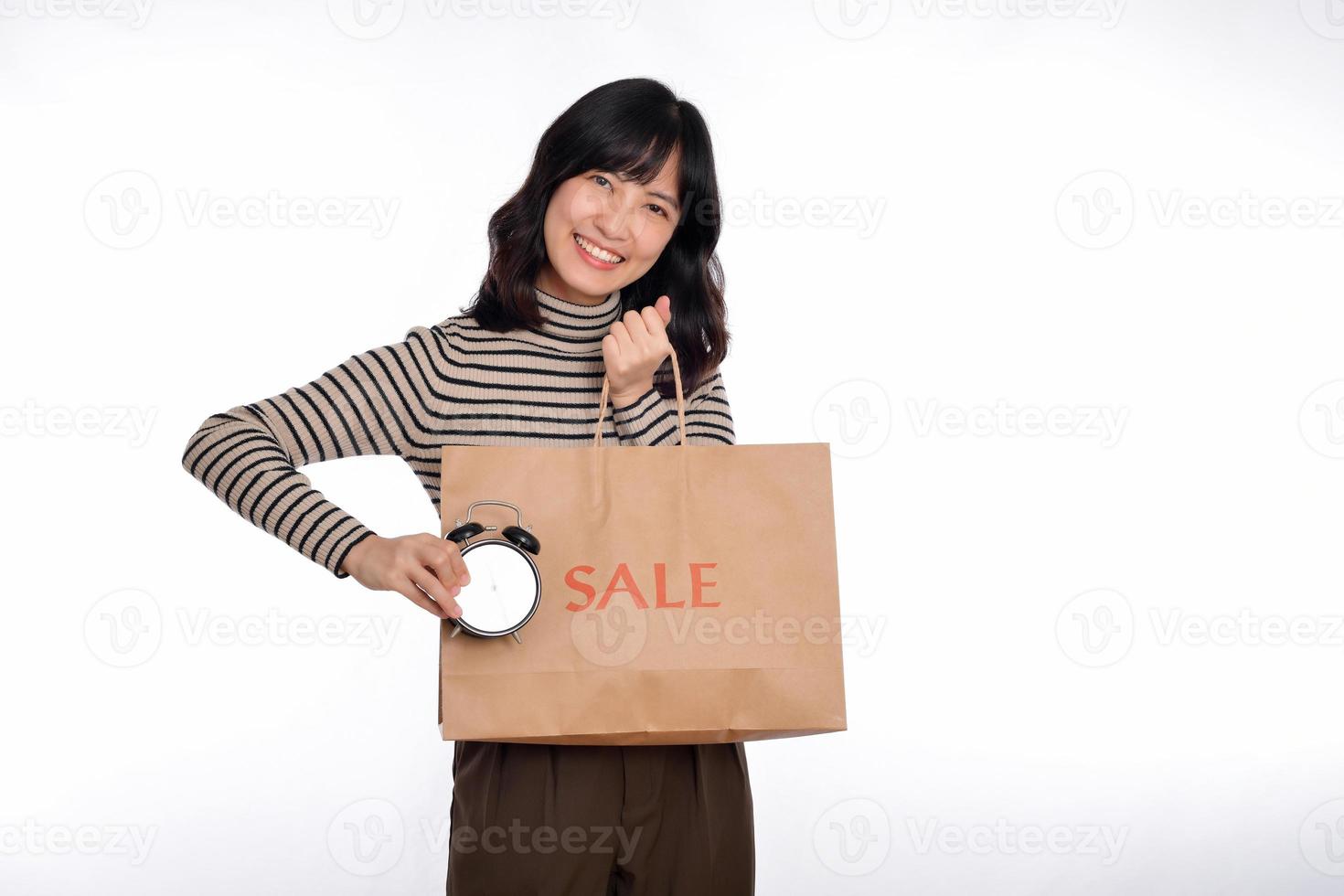 Portrait of happy young Asian woman with sweater shirt holding alarm clock and shopping paper back isolated on white background photo