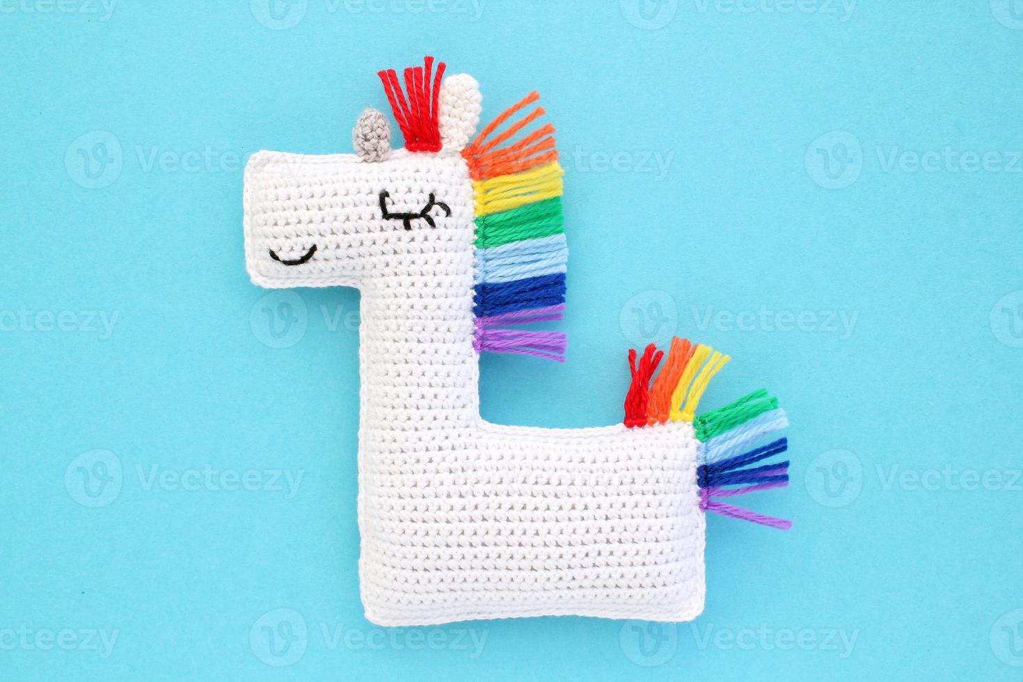 Crochet amigurumi handmade stuffed soft white unicorn toy with rainbow mane on blue background. Handwork, hobby. Craft diy newborn pregnancy concept. Knitted doll for little baby. Close up flat lay photo