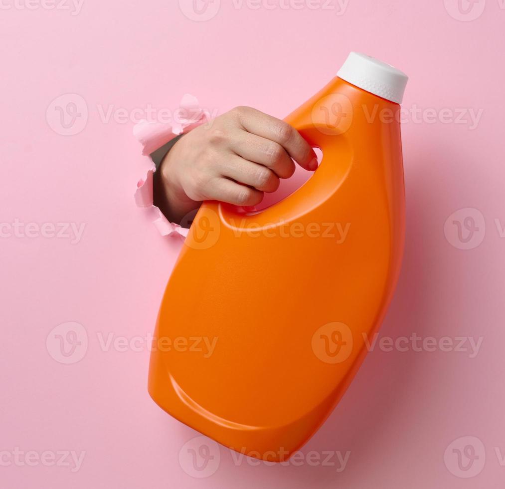 naranja el plastico botella con líquido detergente en un hembra mano en un rosado antecedentes. un parte de el cuerpo palos fuera de un Rasgado agujero en el antecedentes foto