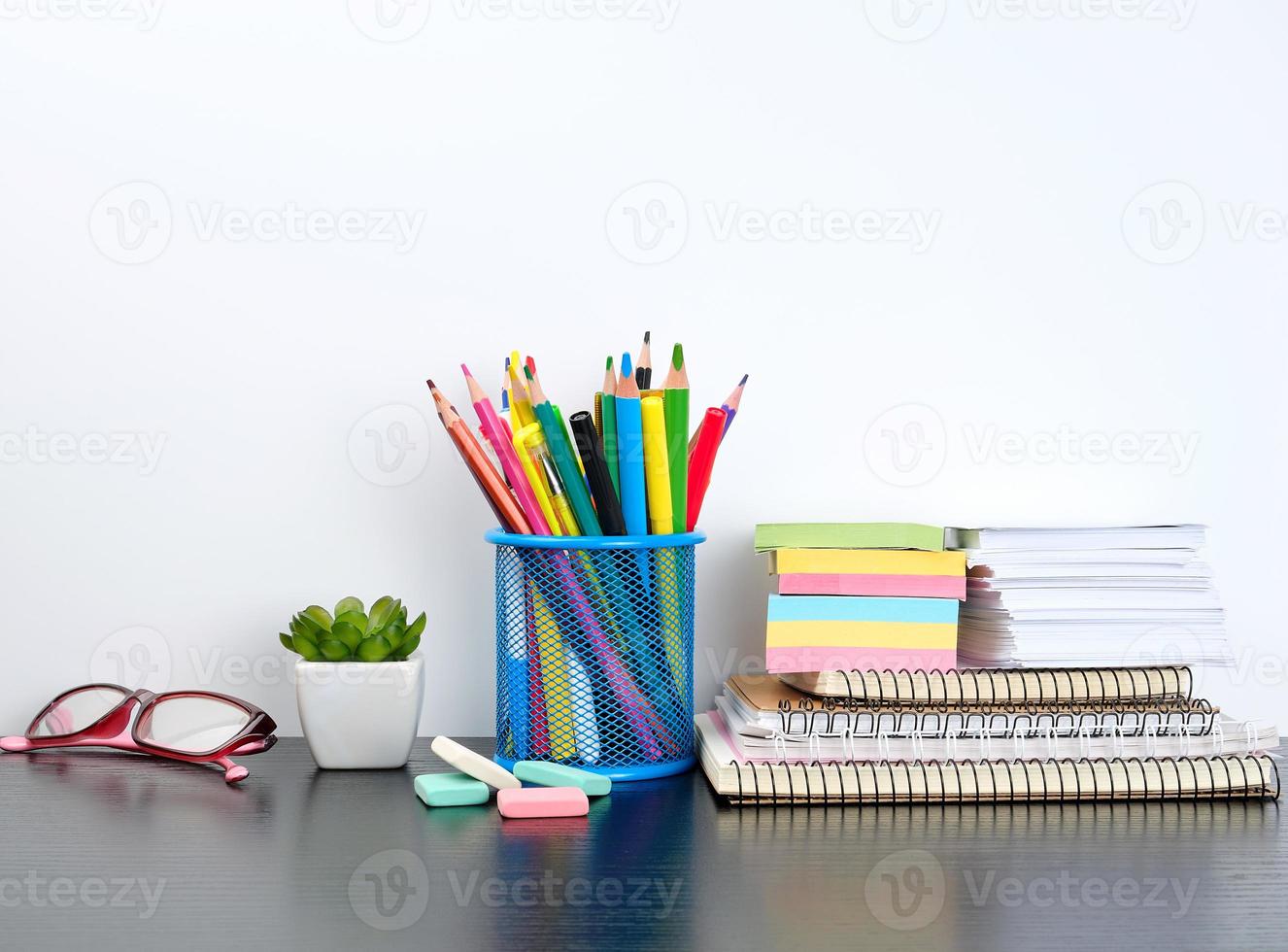 stack of spiral notebooks and colored stickers, next to a ceramic pot with a flower on a black table, white wall photo