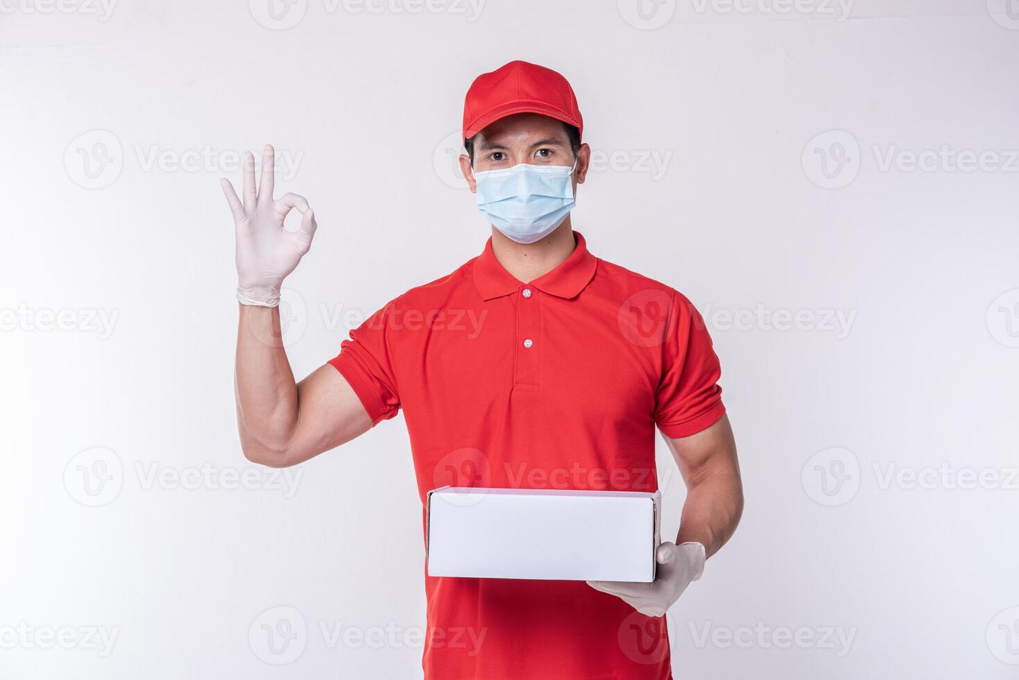 Image of a conscious young delivery man in red cap blank t-shirt uniform face mask gloves standing with empty white cardboard box isolated on light gray background studio photo