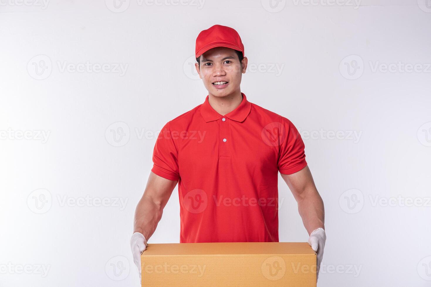 imagen de un joven y feliz repartidor con gorra roja, camiseta en blanco, uniforme de pie con una caja de cartón marrón vacía aislada en un estudio de fondo gris claro foto