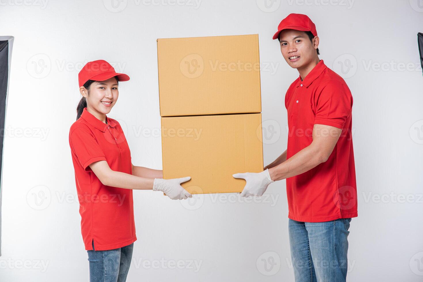 Image of a happy young delivery man in red cap blank t-shirt uniform standing with empty white cardboard box isolated on light gray background studio photo