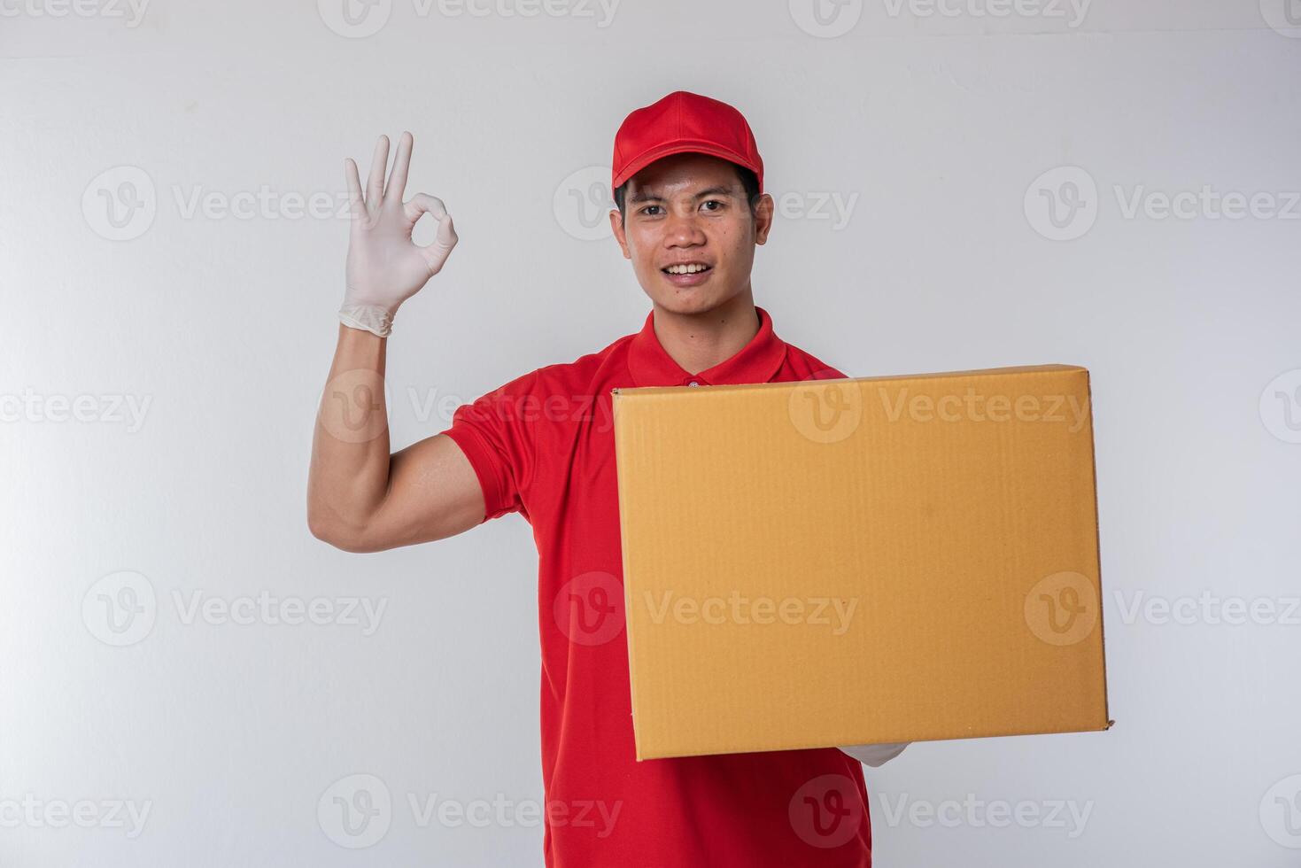 Image of a happy young delivery man in red cap blank t-shirt uniform standing with empty brown cardboard box isolated on light gray background studio photo