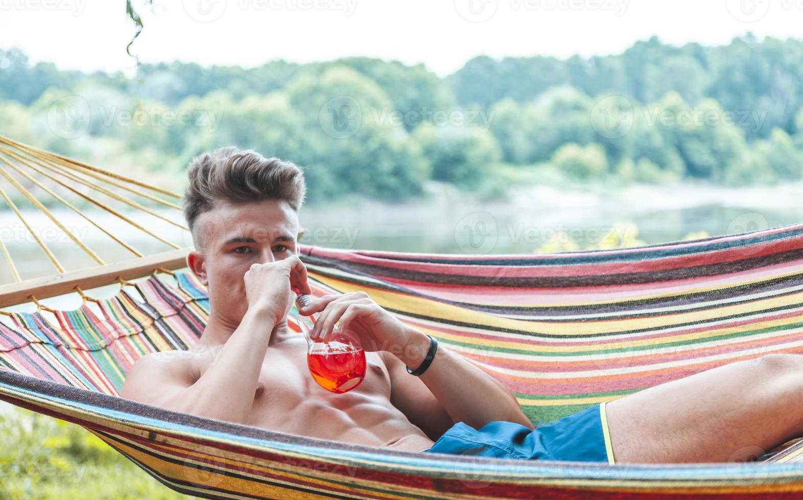 young muscular adult drinking a red drink lying in a hammock photo