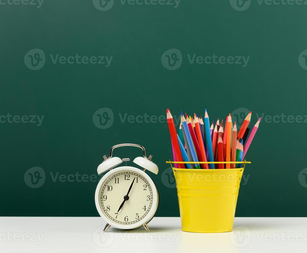 round alarm clock and multicolored pencils on the background of an empty green chalk board. Back to school photo