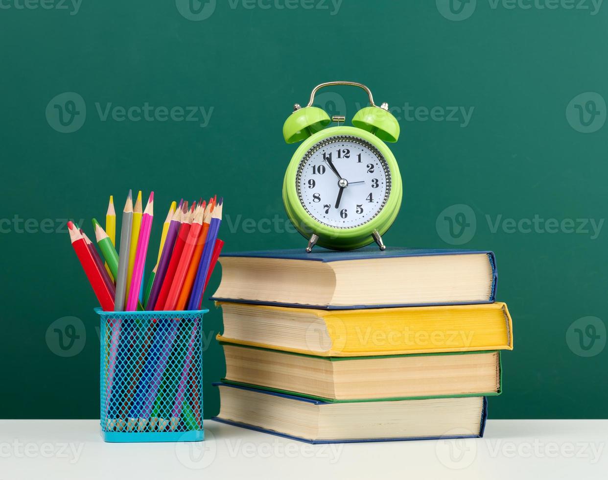 stack of books, a round alarm clock and multicolored pencils on the background of an empty green chalk board photo