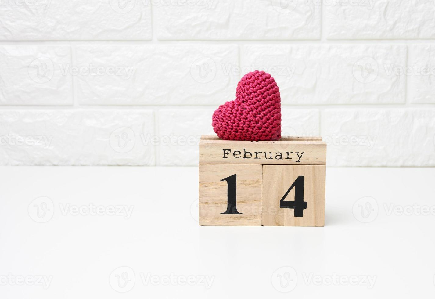 wooden calendar with date February 14 and red knitted heart, white table photo